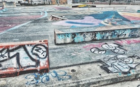 An urban skate park area featuring extensive graffiti art on the concrete surfaces, ramps, and barriers. The ground shows colorful murals and abstract designs, with a mix of spray-painted tags and drawings. Concrete barriers and ramps create a dynamic urban environment.