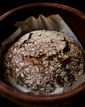 A rustic loaf of bread with a crusty, patterned top sits in a round baking dish lined with parchment paper. The loaf has a golden-brown color with a flour-dusted surface, showcasing a homemade, artisanal appearance.