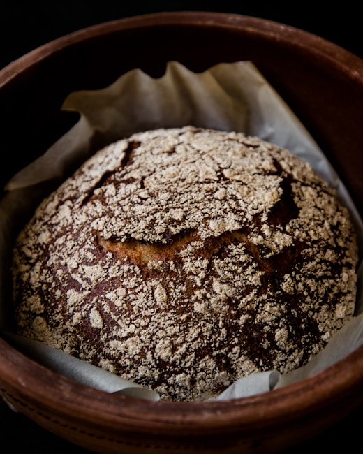 A rustic loaf of bread with a crusty, patterned top sits in a round baking dish lined with parchment paper. The loaf has a golden-brown color with a flour-dusted surface, showcasing a homemade, artisanal appearance.