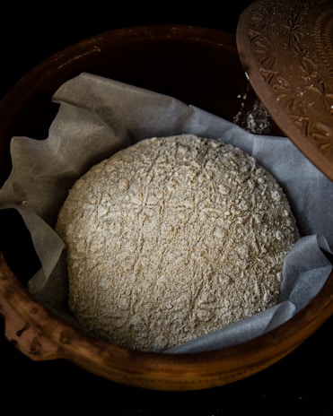 A rustic, unbaked loaf of bread dusted with flour rests on a layer of parchment paper inside a decorative clay bowl with a patterned lid partially opened.