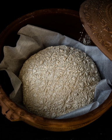 A rustic, unbaked loaf of bread dusted with flour rests on a layer of parchment paper inside a decorative clay bowl with a patterned lid partially opened.
