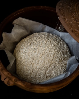 A rustic, unbaked loaf of bread dusted with flour rests on a layer of parchment paper inside a decorative clay bowl with a patterned lid partially opened.