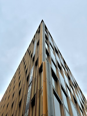 A modern multi-story building with a sharp geometric design, featuring reflective glass windows aligned symmetrically along its facade. The exterior consists of vertical brown and beige panels.
