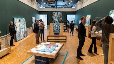 A gallery space with several people interacting with each other, examining various abstract artworks displayed and some pieces still in wooden crates. The room has dark green walls and a wooden parquet floor. The ceilings feature lighting installations.