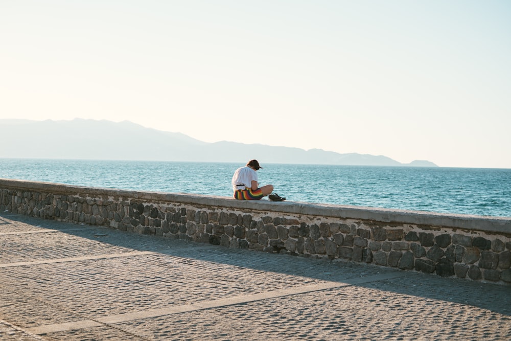 um homem sentado em uma parede de pedra ao lado do oceano