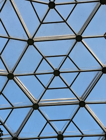 A geometric pattern composed of metal beams forming a honeycomb structure with glass panels. The sky is visible through the glass, providing a backdrop of light blue.