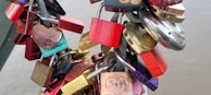 A collection of colorful padlocks attached to a metal chain, often symbolizing love and commitment. Some locks have engravings or names written on them, while various shapes, including heart-shaped locks, are visible. The background is blurred, suggesting these locks are affixed to a structure like a bridge.