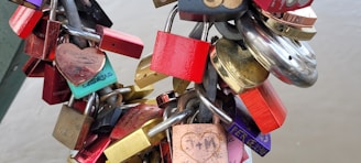 A collection of colorful padlocks attached to a metal chain, often symbolizing love and commitment. Some locks have engravings or names written on them, while various shapes, including heart-shaped locks, are visible. The background is blurred, suggesting these locks are affixed to a structure like a bridge.