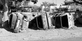 Three overturned cars are lying on their sides next to a concrete wall. The vehicles appear to be old and abandoned, with a dusty and dilapidated appearance. Trees and buildings are visible in the background, casting shadows on the scene.