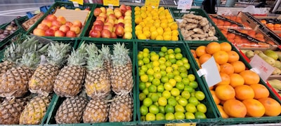 A colorful display of fresh fruits and vegetables at a market. Pineapples are neatly stacked next to green limes and oranges. Nearby, there are apples and other fruits. The arrangement is vibrant and organized, with labels indicating prices and varieties.