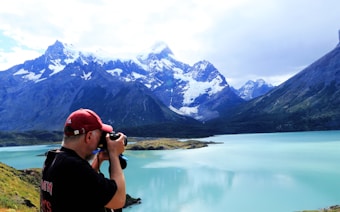 A photographer stands on a grassy area overlooking a scenic view of a large lake with turquoise water, surrounded by towering snow-capped mountains under a partly cloudy sky.