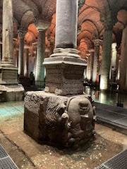 A large ancient stone column stands on a carved stone base, featuring an upside-down Medusa head. The setting is underground, with other columns arranged in rows, creating a sense of depth. The ceiling is constructed with arches of bricks, and the lighting casts a warm glow within the space.
