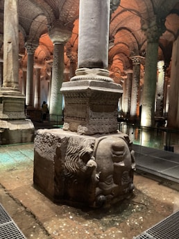A large ancient stone column stands on a carved stone base, featuring an upside-down Medusa head. The setting is underground, with other columns arranged in rows, creating a sense of depth. The ceiling is constructed with arches of bricks, and the lighting casts a warm glow within the space.