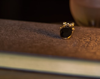 A small piece of ornate jewelry with a dark gemstone and gold setting rests on a textured, possibly aged, surface. Warm lighting casts a soft shadow, enhancing the luxurious and intimate atmosphere.