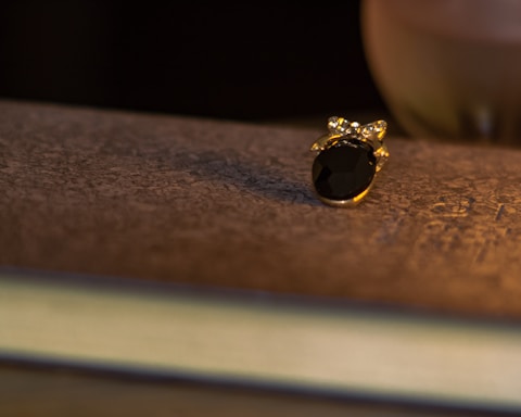 A small piece of ornate jewelry with a dark gemstone and gold setting rests on a textured, possibly aged, surface. Warm lighting casts a soft shadow, enhancing the luxurious and intimate atmosphere.