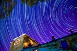 A time-lapse photograph captures star trails in a vibrant night sky above a rustic building. The swirling patterns of light create a sense of motion and wonder. The structure, made of stone with wooden windows, contrasts with the vivid blues and purples of the celestial display.