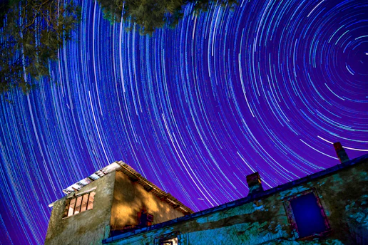 A time-lapse photograph captures star trails in a vibrant night sky above a rustic building. The swirling patterns of light create a sense of motion and wonder. The structure, made of stone with wooden windows, contrasts with the vivid blues and purples of the celestial display.