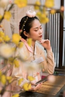 A person in traditional clothing sitting thoughtfully, surrounded by delicate blossoms. Their attire is elegant with soft colors and intricate details. The setting features wooden elements and serene decor, contributing to a calm atmosphere.