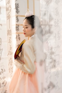 A woman wearing traditional Korean hanbok stands gracefully, holding a colorful fan. Her eyes are closed, and she wears a delicate accessory in her hair. Korean calligraphy adorns the semi-transparent curtain in the background.