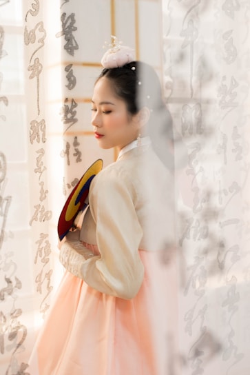 A woman wearing traditional Korean hanbok stands gracefully, holding a colorful fan. Her eyes are closed, and she wears a delicate accessory in her hair. Korean calligraphy adorns the semi-transparent curtain in the background.