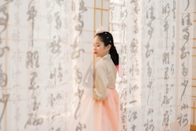 A woman wearing traditional Korean hanbok stands gracefully between translucent curtains that have calligraphy on them. She appears serene and composed, with her eyes closed and hands gently folded in front of her. The lighting is soft and diffused, casting a gentle glow on her face and the fabric around her.