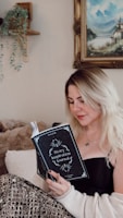 A woman with blonde hair is sitting comfortably and reading a 'Story Inspiration Journal'. She is dressed in a black top and a cozy, patterned blanket drapes over her legs. A soft plush blanket and a wall with a hanging plant and a landscape painting enhance the cozy setting.