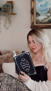 A woman with blonde hair is sitting comfortably and reading a 'Story Inspiration Journal'. She is dressed in a black top and a cozy, patterned blanket drapes over her legs. A soft plush blanket and a wall with a hanging plant and a landscape painting enhance the cozy setting.