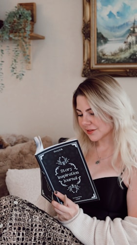 A woman with blonde hair is sitting comfortably and reading a 'Story Inspiration Journal'. She is dressed in a black top and a cozy, patterned blanket drapes over her legs. A soft plush blanket and a wall with a hanging plant and a landscape painting enhance the cozy setting.