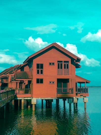 A stilted wooden house stands over calm water, with a bright sky and scattered white clouds. The house has multiple small windows and is supported by wooden pillars, with a rooftop terrace visible on one side.