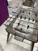 A modern foosball table featuring transparent glass surfaces and polished metal rods. The players are sculpted in light and dark wood tones with intricate handles. The tabletop design is sleek, with a combination of minimalist and rustic elements.