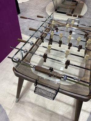 A modern foosball table featuring transparent glass surfaces and polished metal rods. The players are sculpted in light and dark wood tones with intricate handles. The tabletop design is sleek, with a combination of minimalist and rustic elements.