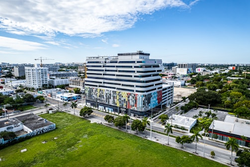 A modern, multi-story building with a colorful facade stands prominently in an urban setting, surrounded by streets lined with palm trees. Other commercial and residential buildings can be seen in the background, along with a construction crane. The sky is partly cloudy, adding to the vibrant cityscape.