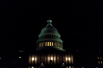 A well-lit, iconic dome of a capitol building stands illuminated against a dark night sky. The warm glow of lights from within the building creates a stark contrast with the deep shadows surrounding it.
