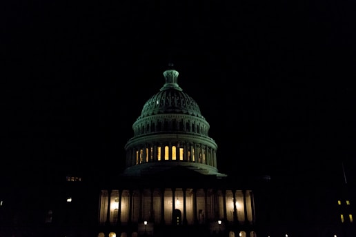 A well-lit, iconic dome of a capitol building stands illuminated against a dark night sky. The warm glow of lights from within the building creates a stark contrast with the deep shadows surrounding it.