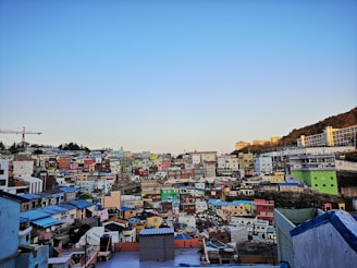 A densely packed hillside neighborhood of colorful houses under a clear blue sky during daylight. The houses are painted in various bright colors including green, yellow, pink, and blue, creating a vibrant mosaic effect. There is a construction crane visible on the left, suggesting ongoing development. In the background, hills and a modern structure can be seen.