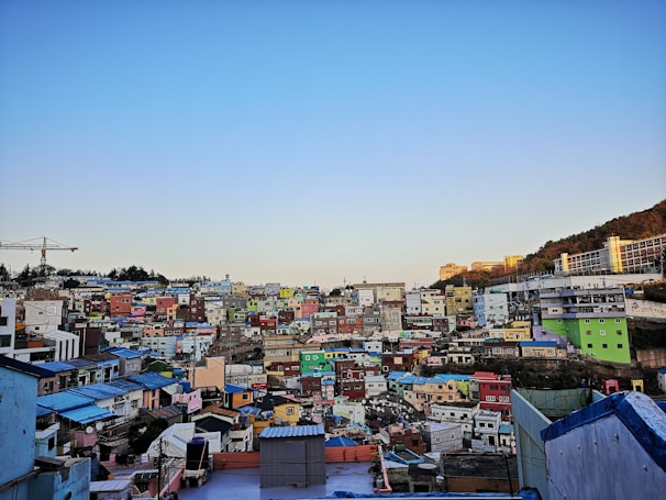 A densely packed hillside neighborhood of colorful houses under a clear blue sky during daylight. The houses are painted in various bright colors including green, yellow, pink, and blue, creating a vibrant mosaic effect. There is a construction crane visible on the left, suggesting ongoing development. In the background, hills and a modern structure can be seen.