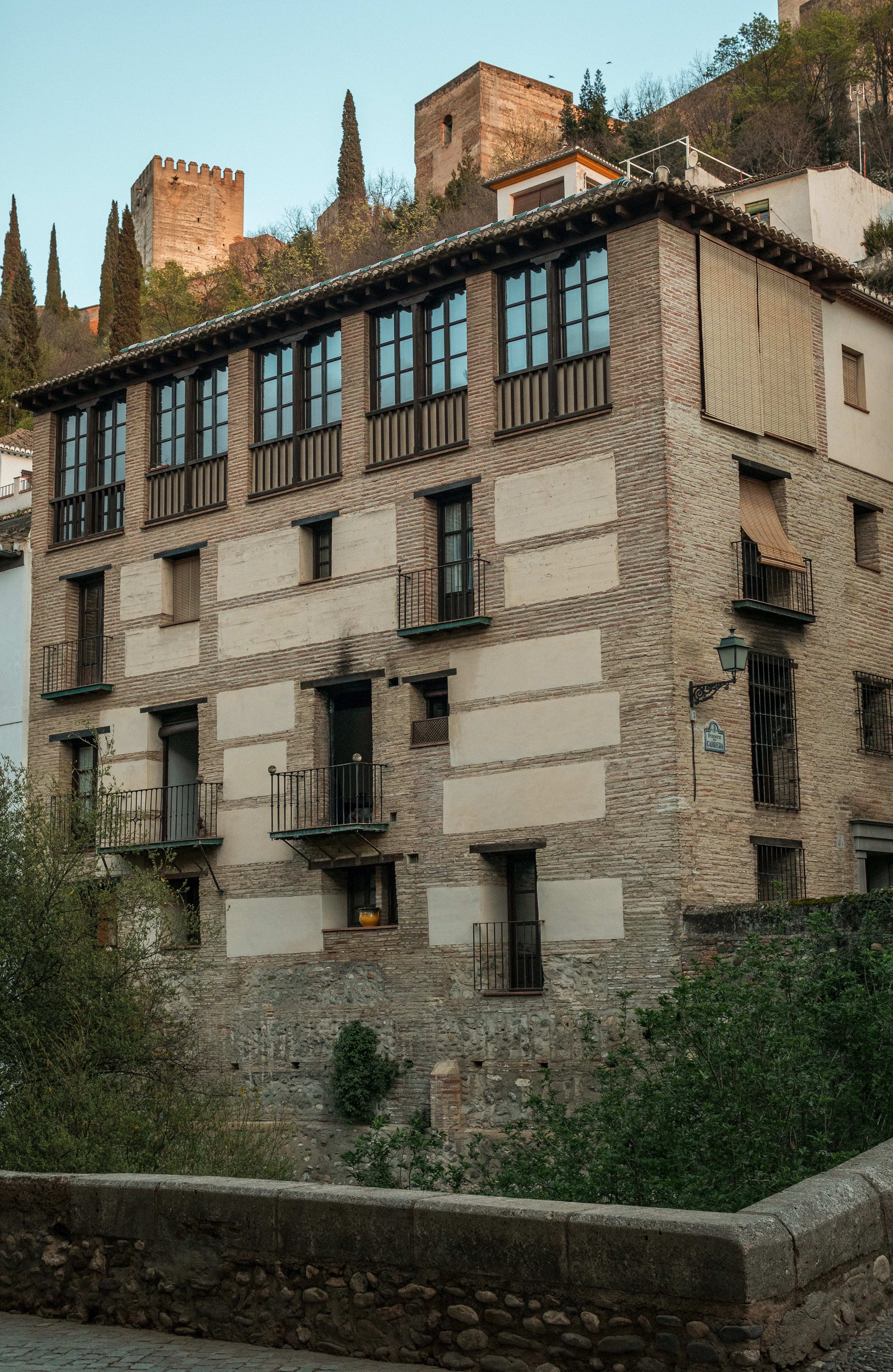 A tall historic Granada house of brick and stone banding with a glazed timber gallery running the length of its top floor and small iron balconies below, a medieval tower on the wooded hill above.