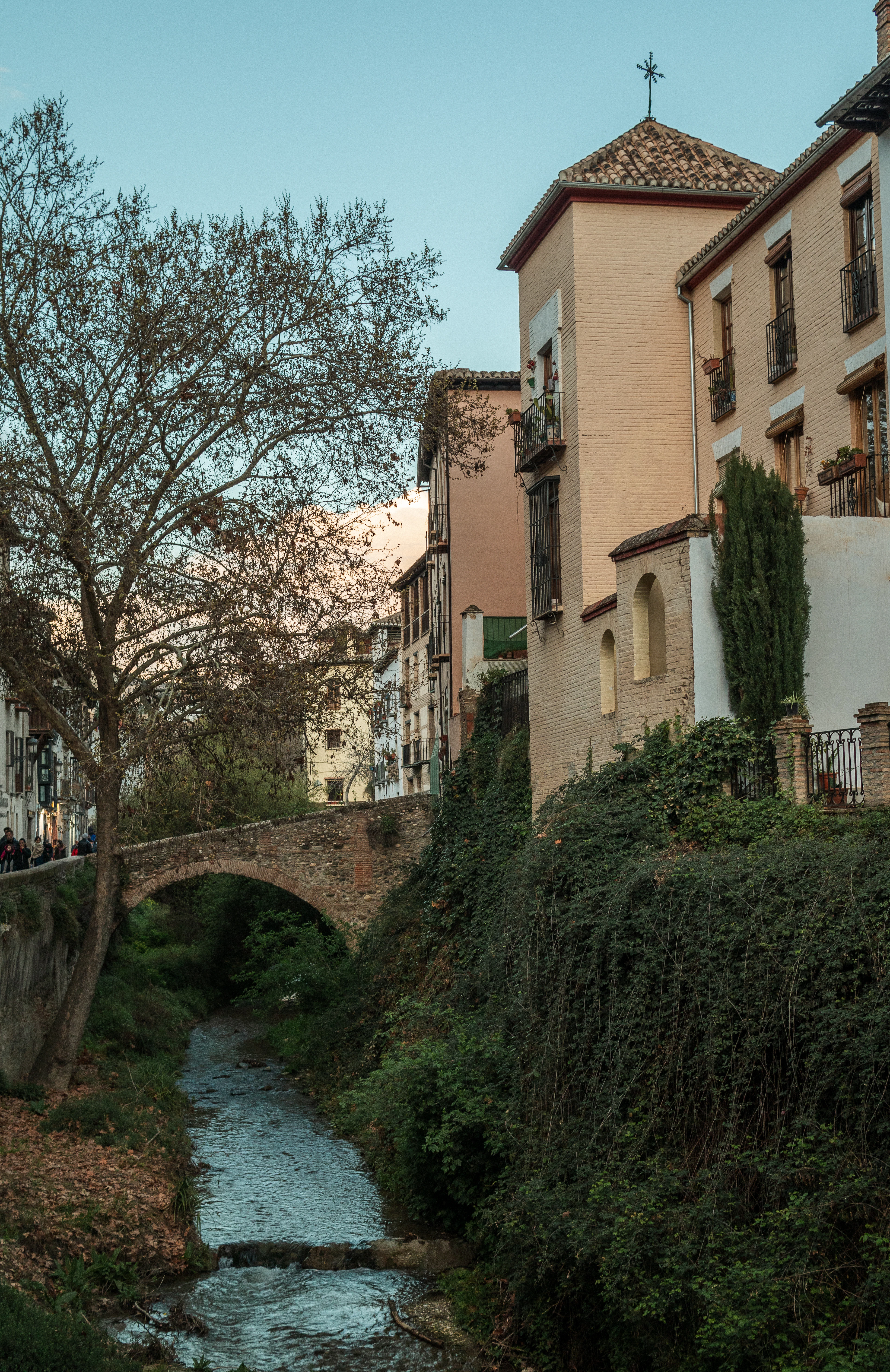 The narrow Darro river running beneath a small stone arch bridge in Granada at dusk, ivy-covered banks below tiled-roof houses with wrought-iron balconies and a slim cypress.
