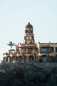 A large, luxurious villa with Mediterranean architectural style, featuring arched windows and balconies. The building has multiple levels and a central tower with a dome. Palm trees are situated around the structure, and it is located on a rocky cliff, suggesting a coastal setting.