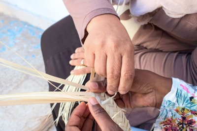 Several hands are involved in the intricate task of weaving strips of natural fiber, showcasing a collaborative effort in a craft activity. The background is blurred, focusing attention on the hands and the weaving process.