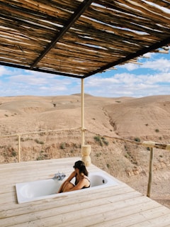 A person relaxes in an outdoor bathtub on a wooden deck overlooking a rugged desert landscape. Natural wooden beams form a partial roof, providing some shade. The sky is partially cloudy, and the terrain around is arid and expansive.