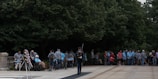 A ceremonial guard in a uniform stands on a paved area surrounded by numerous people who appear to be observing an event. Many of the observers are older individuals, some in wheelchairs, suggesting a possible veterans' gathering. The background is dominated by lush green trees, creating a serene atmosphere. A wreath stands to the left of the guard, adding a ceremonial or memorial aspect to the scene.