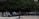 A ceremonial guard in a uniform stands on a paved area surrounded by numerous people who appear to be observing an event. Many of the observers are older individuals, some in wheelchairs, suggesting a possible veterans' gathering. The background is dominated by lush green trees, creating a serene atmosphere. A wreath stands to the left of the guard, adding a ceremonial or memorial aspect to the scene.