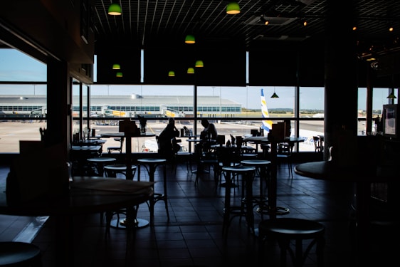 Dimly lit airport café with silhouetted tables and chairs. Large windows reveal a view of the runway and an airplane parked outside.