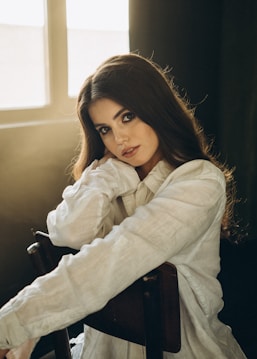 A woman with long brown hair wears a flowing white blouse and sits on a wooden chair next to a softly lit window. The warm light bathes her face, creating a serene and introspective atmosphere. She gazes directly at the camera with a calm expression and rests her chin on her hand, conveying a sense of elegance and poise.