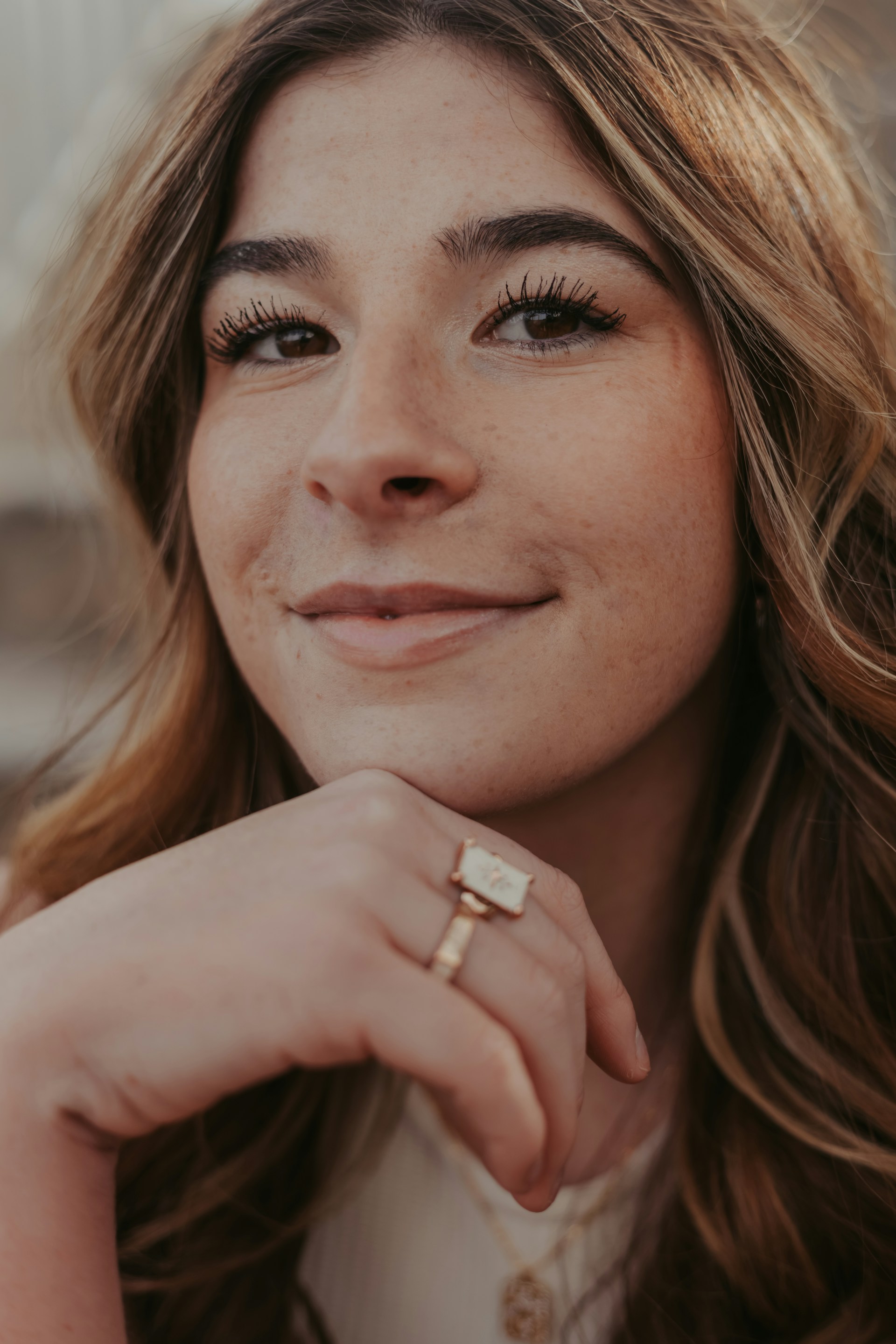 A person with long, wavy hair gazes confidently at the camera with a slight smile. They have prominent eyelashes and wear a ring on their finger. The lighting is soft and warm, accentuating their facial features.