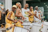A group of people are energetically playing drums possibly in a street parade or festival. They are dressed in brightly colored, coordinated outfits. Others in the background appear to be similarly dressed, creating a lively and engaging atmosphere.