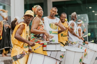 A group of people are energetically playing drums possibly in a street parade or festival. They are dressed in brightly colored, coordinated outfits. Others in the background appear to be similarly dressed, creating a lively and engaging atmosphere.