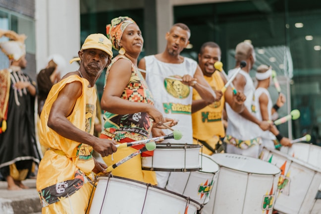 A group of people are energetically playing drums possibly in a street parade or festival. They are dressed in brightly colored, coordinated outfits. Others in the background appear to be similarly dressed, creating a lively and engaging atmosphere.