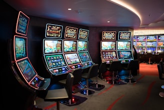 A row of brightly lit slot machines in a casino setting, each with colorful displays and various game themes. The machines are lined up against a dark wall and are unoccupied, with cushioned chairs placed in front of them. The area is dimly lit except for the glow from the screens, creating a vibrant and engaging atmosphere.
